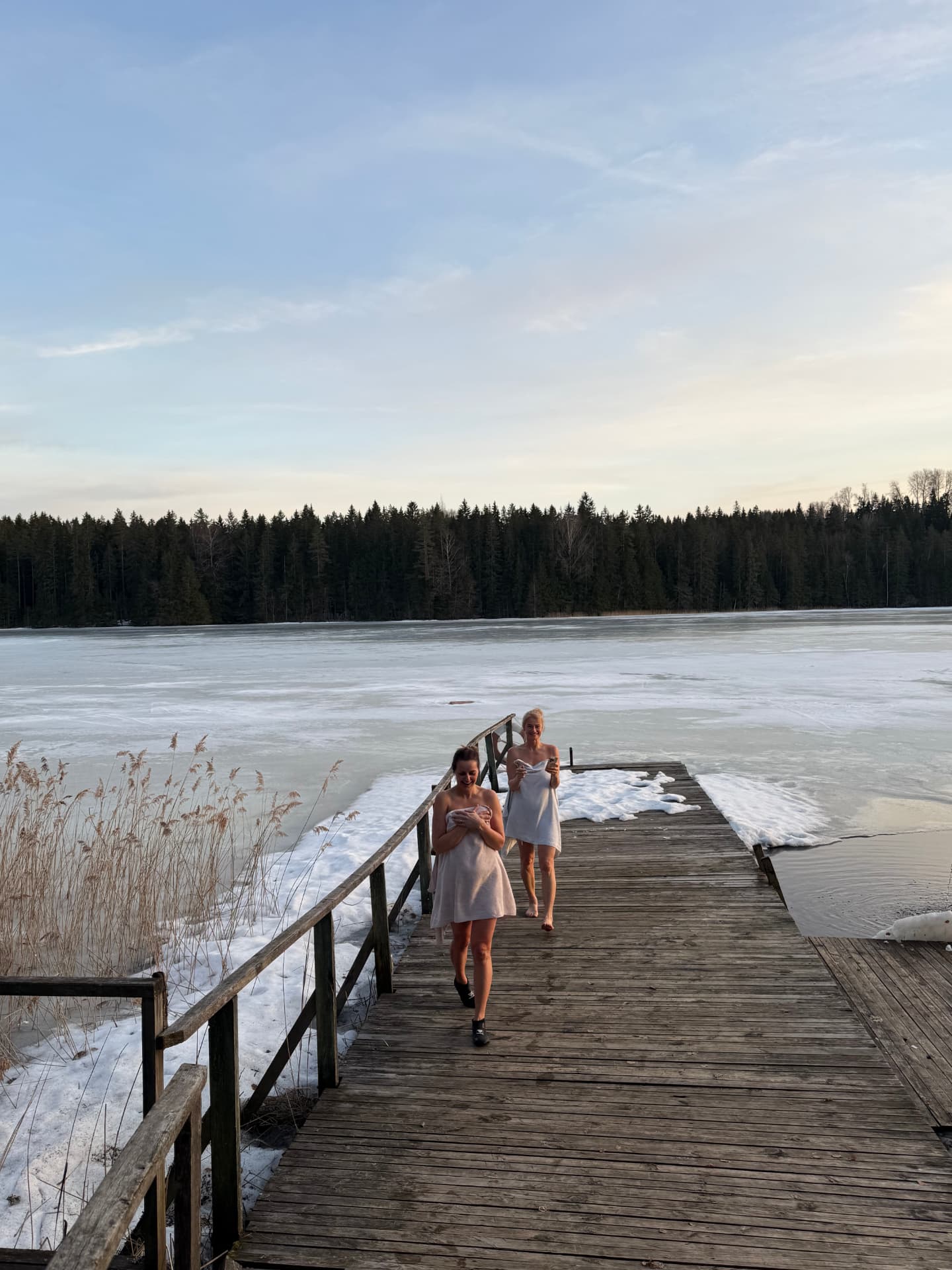 Kaks naist puidust kail pärast sauna, järv osaliselt jääs, mets taustal